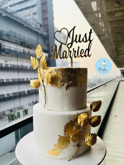 An elegant two-tier 'Just Married' cake in white and gold. This sophisticated design features a white fondant base with gold leaf accents and delicate branches with golden leaves, perfect for a modern wedding.