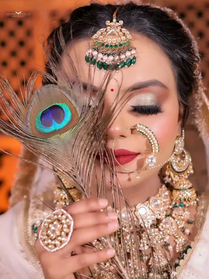 An artistic portrait where the bride holds a peacock feather to her face. The vibrant blue and green of the feather contrasts beautifully with her white and gold attire.