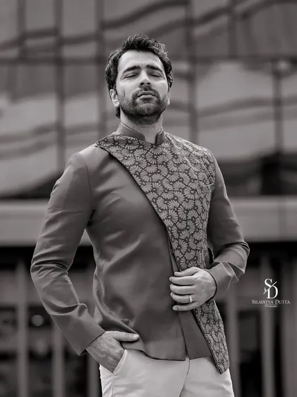 A black and white portrait of Abir Chatterjee against a modern architectural background. The strong lines of the building complement the sharp tailoring of his outfit.