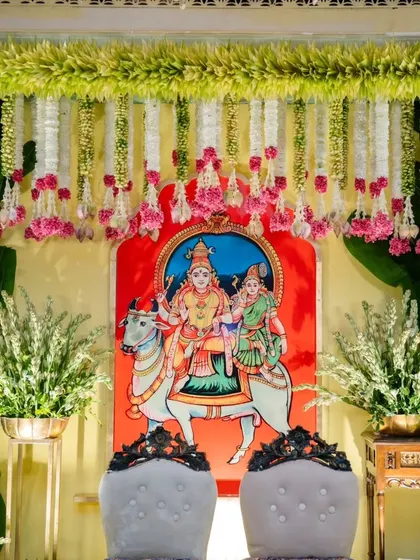 The backdrop for a Muhurtham ceremony, featuring a traditional painting of deities. The scene is framed with beautiful tuberose and pink rose garlands, blending art with floral design.