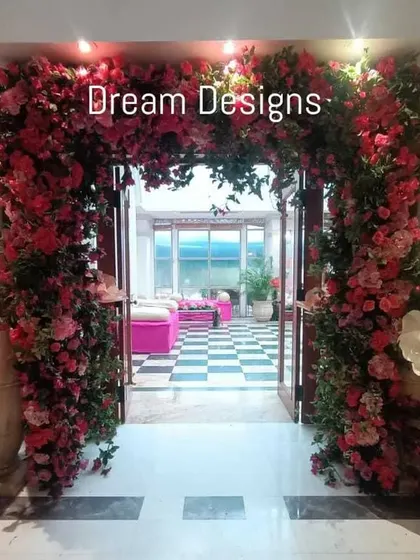 A grand floral entrance designed for a Mehendi party. The doorway is framed by a massive arch of pink flowers and oversized paper blooms, creating a vibrant and joyful welcome.