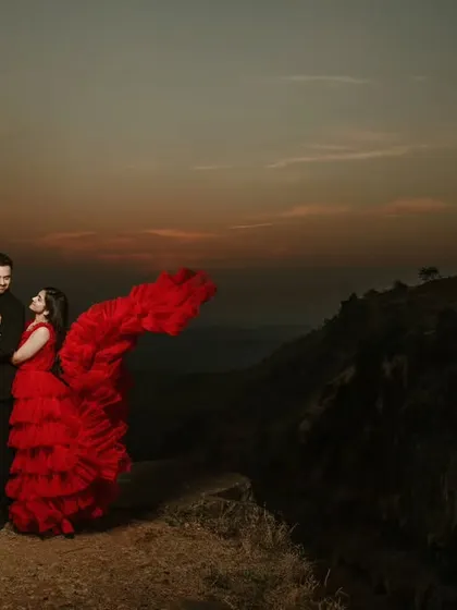 A wide-angle shot capturing the grandeur of a mountain pre-wedding location. The bride's ruffled red dress adds a pop of color to the dusky, atmospheric landscape.