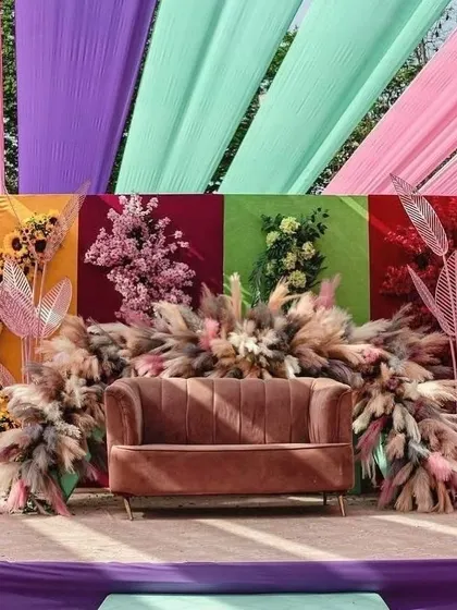 A colorful stage backdrop for a Mehendi or Haldi function. The design uses panels of different colors, each with a unique floral arrangement, and a central explosion of pampas grass.
