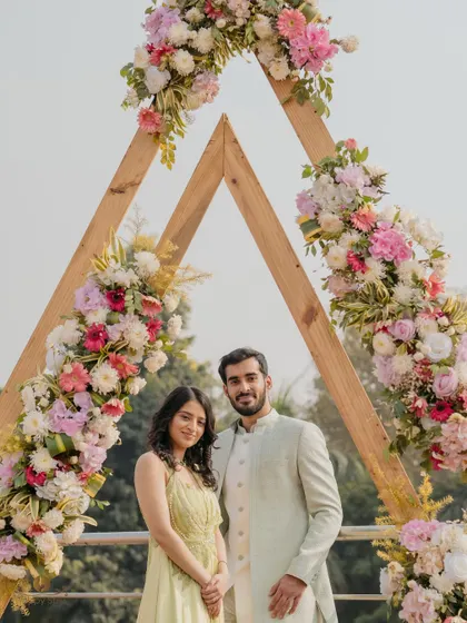 A lovely portrait of the couple standing under a beautiful floral arch at their roka ceremony. The pastel colors of their outfits and the decor create a soft and romantic atmosphere.