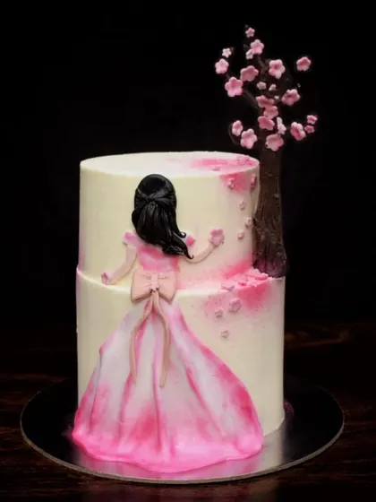 A whimsical and dreamy two-tier cake. It features a hand-painted fondant figure in a flowing pink gown, standing next to a delicate cherry blossom tree.