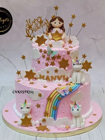 A dreamy two-tier pink cake for a birthday celebration. It's adorned with a fondant girl holding a star, cute unicorns, a rainbow slide, and golden star accents.