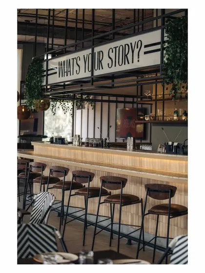 A side view of the bar, highlighting the clean lines of the bar stools and the prominent placement of the brand's tagline. The industrial grid above frames the entire scene.