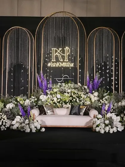 A modern and elegant photo-op stage for a Sangeet, featuring clean golden arches, cascading crystal strings, and a lush bed of white and purple florals.