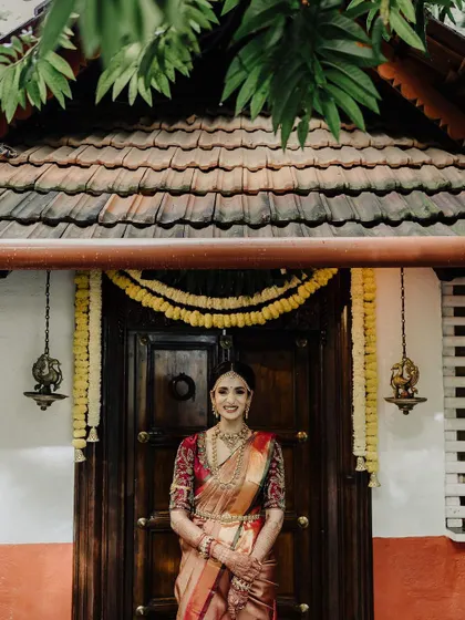 A timeless bridal portrait set against a traditional South Indian architectural backdrop. The bride's graceful pose and elegant saree are the centerpiece of this beautiful shot.