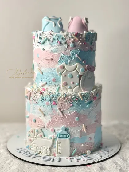A "He or She" gender reveal cake with a textured pink and blue finish. It's decorated with cute fondant baby items like onesies, a bottle, and a carriage.