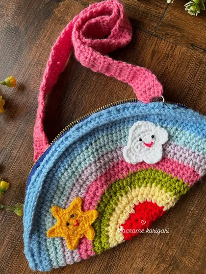 Our handmade rainbow crochet bag for kids is designed to bring a smile to any child's face. It features a colorful rainbow arc, cute star and cloud appliqués, and a bright pink strap.