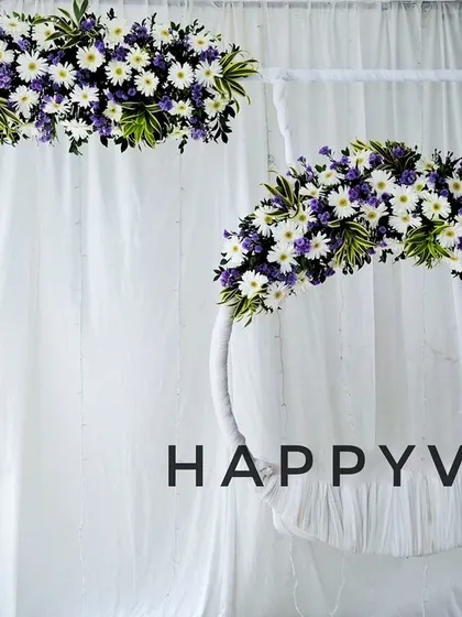 A clean and elegant backdrop design featuring white drapes and floral arrangements of white daisies and purple chrysanthemums. The hanging floral hoop adds a simple yet beautiful touch for any celebration.