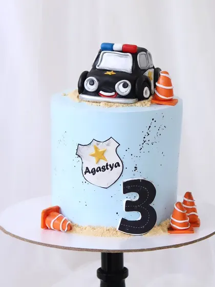 A cute police car cake for a third birthday. This light blue cake features a cartoonish, handcrafted police car topper, complete with traffic cones and a personalized badge.