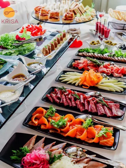 A long, elegant table arrangement featuring a variety of cured meats like salami and prosciutto, smoked salmon, and fresh salads. The linear setup is perfect for allowing guests to move along and sample different items easily.