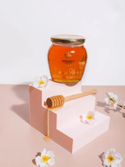 A minimalist and elegant shot of honey. The product is placed on a pink staircase prop, with a delicate honey drip and white flowers adding a touch of sophistication.