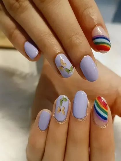 A playful and artistic set combining pastel purple nails with butterfly decals and rainbow-striped accent nails.