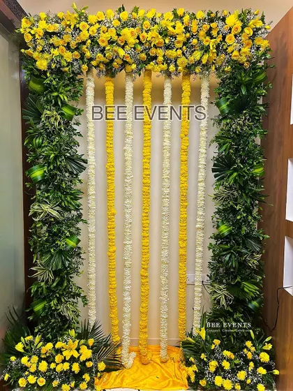 A simple and elegant Haldi backdrop perfect for an indoor celebration. The design features a frame of lush greenery and yellow flowers, with hanging garlands of tuberose and marigold.