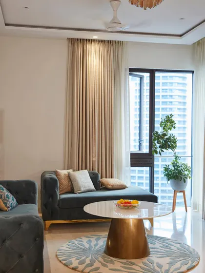 A cozy seating nook in a Lodha apartment, featuring a deep blue chaise lounge and a modern coffee table, with the city view as a stunning backdrop.