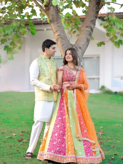 A classic pre-wedding portrait. The couple stands together in a garden setting, their traditional outfits and happy expressions perfectly captured.