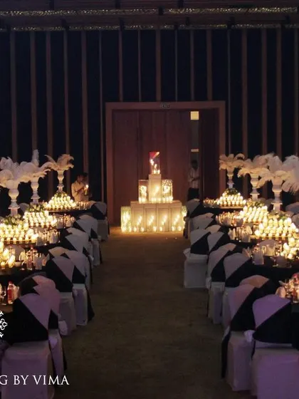 A glamorous anniversary celebration with a dramatic red and black theme. The room is filled with candlelit tables, large white feather centerpieces, and a stunning stage with a heart-shaped light installation.