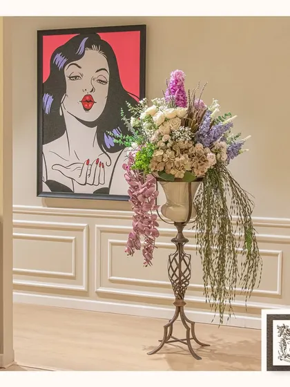 This lush artificial floral arrangement brings nature indoors in the most enchanting way. Placed on a tall metal pedestal, it adds a dramatic and elegant touch to this corner, complemented by playful pop art.