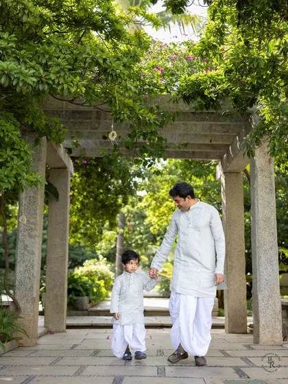 A father and son, dressed in matching white kurtas and dhotis, walk hand-in-hand through a beautiful garden path.