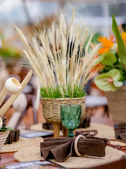 A centerpiece of dried wheat stalks in a woven basket, set next to a green glass goblet. The decor celebrates natural textures and materials.