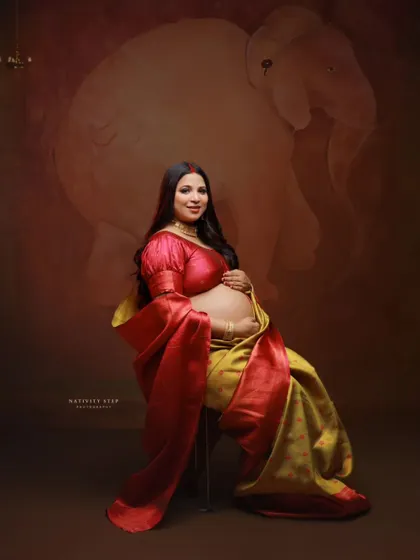 A solo portrait of the mother-to-be seated, showcasing her baby bump in a beautiful yellow and red saree. The pose is both regal and gentle.