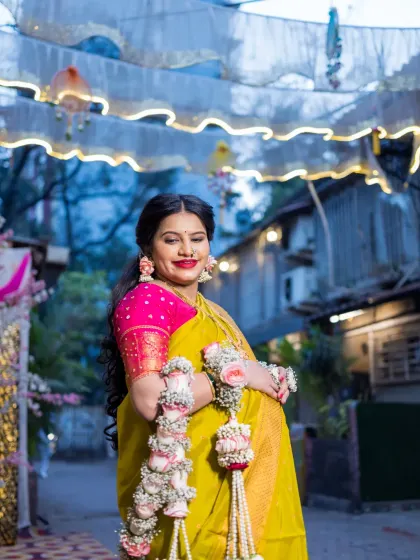 The mother-to-be smiles radiantly, standing under festive outdoor decorations for her baby shower. This shot captures her joy and the celebratory atmosphere of the event, highlighting her traditional attire and floral ornaments.