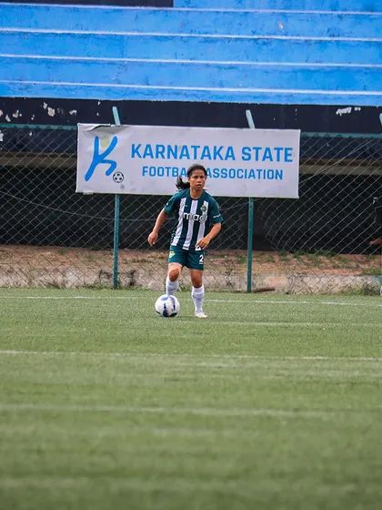 A player stands ready with the ball at her feet, positioned under the Karnataka State Football Association banner at the stadium.