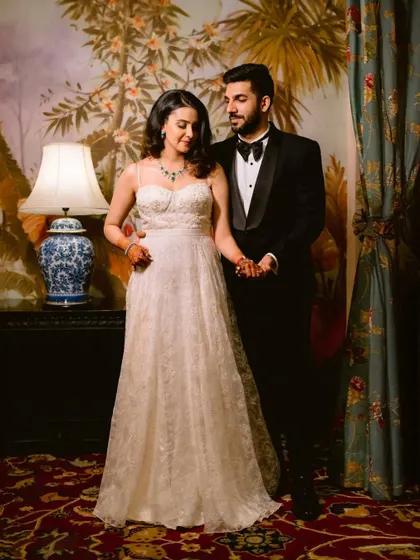 A full-length portrait of the couple, her in a beautiful white gown and him in a classic tuxedo, standing in a lavishly decorated room, exuding timeless elegance.