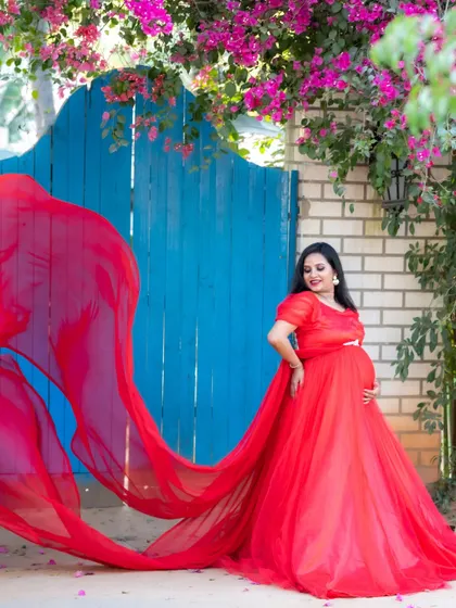 A dramatic shot of a mom-to-be in a red gown against a bright blue gate with pink flowers.