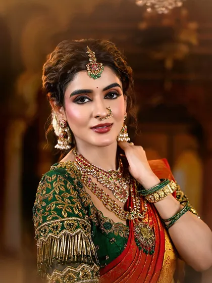 A regal and traditional bridal look featuring a rich red and green Kanjivaram saree. The makeup is defined by warm, shimmery eyes and a deep red lip, perfectly accessorized with temple jewelry.