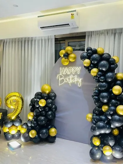 A chic 29th birthday setup at home, with a black and gold balloon arch, a 'Happy Birthday' neon sign, and large foil number balloons.