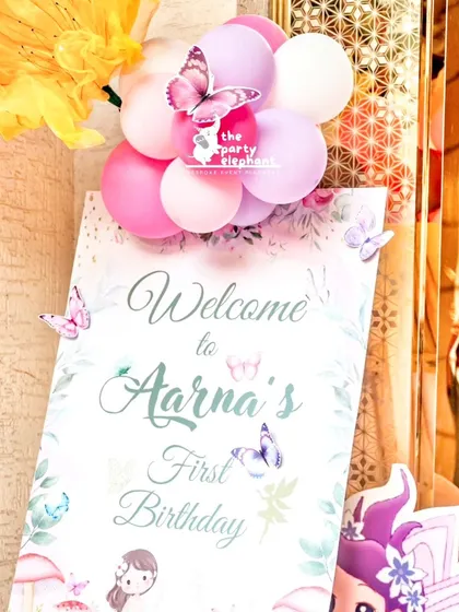 A close-up of the personalized welcome sign for Aarṇa's first birthday, adorned with a small cluster of pink and lilac balloons and a butterfly accent.