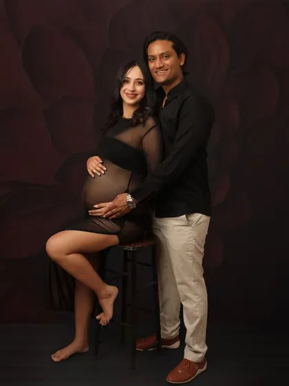 A classic and elegant studio portrait. The couple is dressed in black, posing against a dark, textured backdrop, which creates a sophisticated and intimate mood for their maternity session.