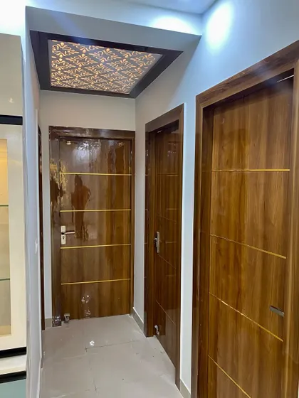 A hallway view showing multiple flush doors with a high-gloss wood finish and gold inlay. The foyer ceiling features a beautiful backlit jali panel.