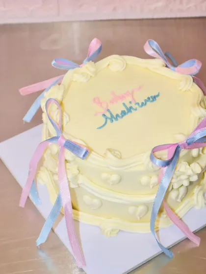 We love the delicate, vintage coquette cake trend. This beautiful baby shower cake features soft yellow buttercream, intricate piping, and is adorned with delicate pink and blue ribbons, making it a perfect centerpiece.
