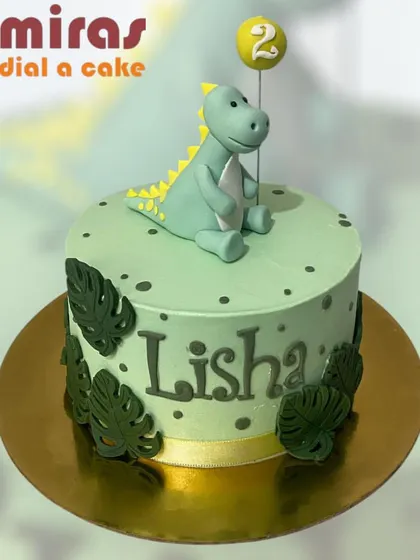 A dino-mite cake for a second birthday! This adorable design features a friendly green dinosaur, jungle leaves, and a balloon topper, all on a pastel green cake that's perfect for a young dinosaur fan.