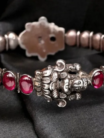 A close-up of the handcrafted silver bracelet from the red stone set. The centerpiece features a finely detailed carving of the goddess Lakshmi, flanked by red stones.