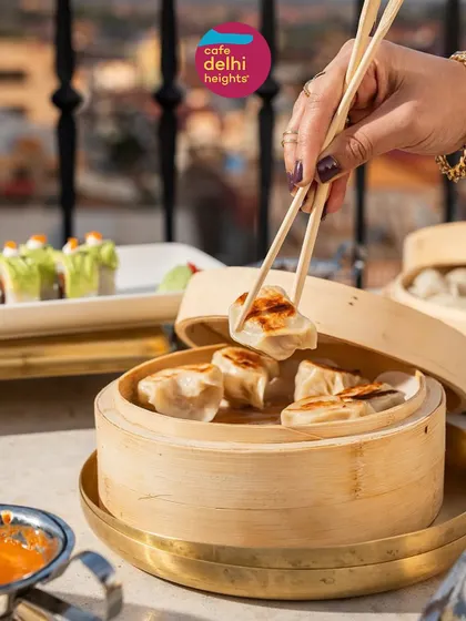 A hand using chopsticks to pick up a pan-fried dumpling from a bamboo steamer at Cafe Delhi Heights. This shows the variety of Asian-inspired dishes available across my different restaurants.