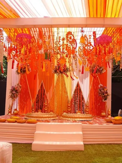 The main stage for the Haldi ceremony, designed for beautiful visuals and fond memories. We used layers of orange and yellow drapes, a backdrop of traditional hangings, and comfortable seating for the couple.
