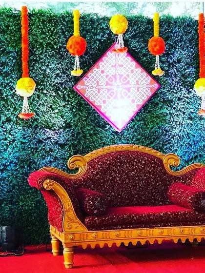 A rich backdrop combining a green grass wall with hanging marigold strings and traditional brass lamps (Samai). The ornate red sofa provides a royal seating area for the main guests.