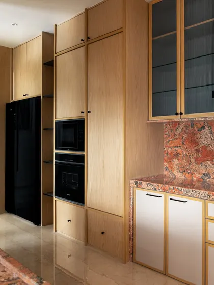 The kitchen in 'ARAH' features light wood cabinetry and a striking red-orange marble backsplash. The tall unit integrates the oven and microwave, creating a seamless and functional design.