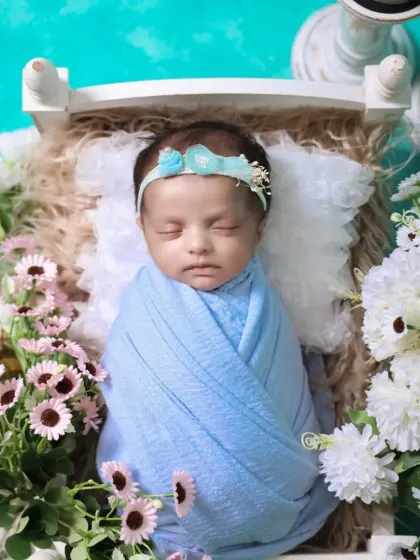 An overhead portrait of a newborn swaddled in blue. The floral headband and surrounding flowers add a soft, delicate touch to this beautiful baby photo.