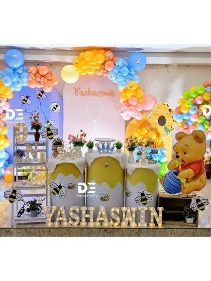 A sweet Winnie the Pooh and honeybee theme for Yashaswin's first birthday. The setup features Pooh Bear with his honey pot, beehive-themed pedestals, and a colorful balloon arch.