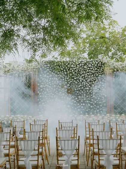An elegant outdoor engagement setup with a wall of white flowers and dramatic smoke effects for the couple's entrance.