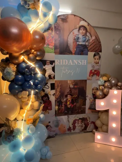 A full view of Ridansh's first birthday setup, showcasing a custom backdrop with his photos, a light-up '1', and a beautiful balloon garland.