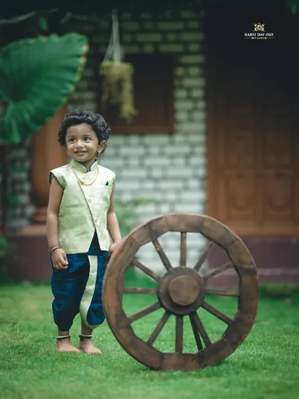 A handsome little boy in a traditional outfit, standing proudly next to a large wooden wheel in our garden.