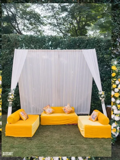 A simple yet elegant Haldi seating area with a bright yellow sofa set against a backdrop of white drapes and lush greenery.